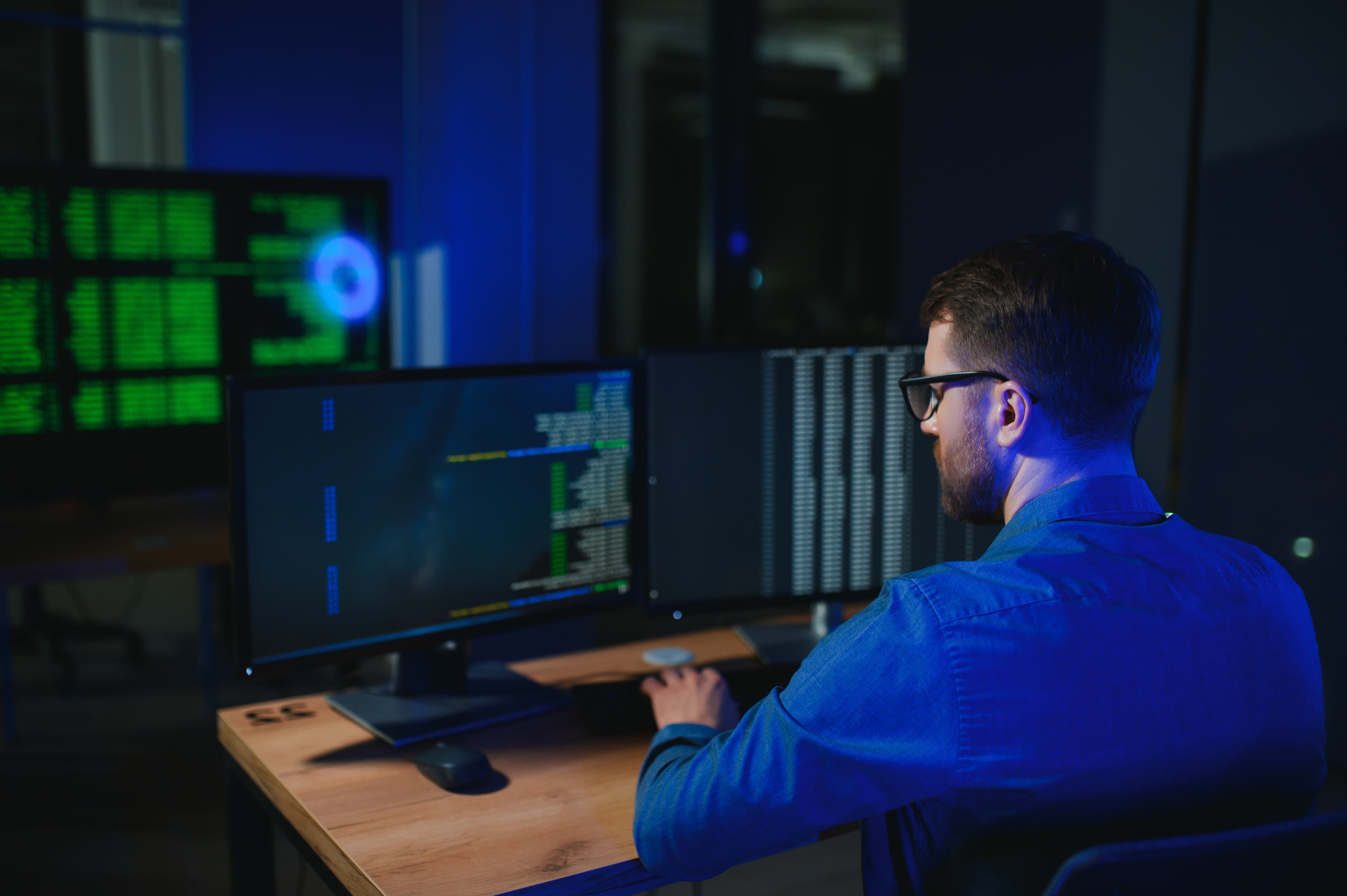 IT security professional monitoring multiple screens of network data at a dark workstation
